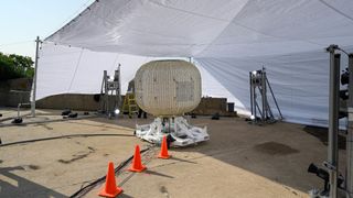 An off-white, almost tan circular inflatable module from Lockheed Martin sits on a test stand like a squished sphere. Three cones line the path of black wires laid on the concrete ground from the module to the foreground. A white canopy hangs over the module, and covers the top half of the image.