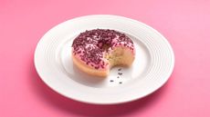 Pink doughnut on white plate on pink background with bite removed