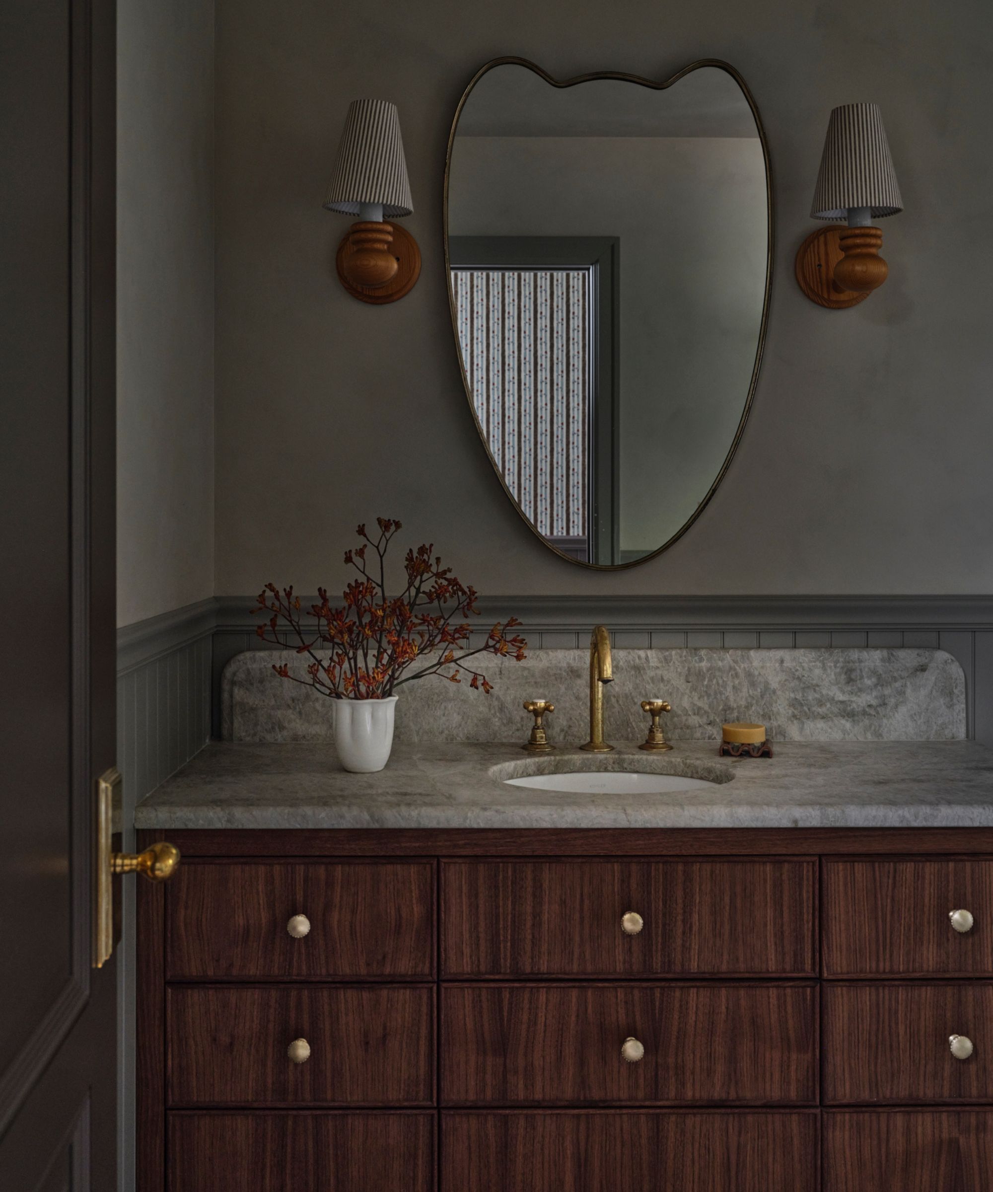 a cottage bathroom vanity area with dark brown wooden vanity, stone counter and a curved mirror with two wooden sconces either side