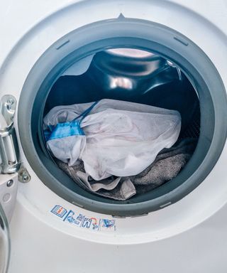 A white washing machine with the door open and the silver drum filled with a white laundry bag and a gray towel