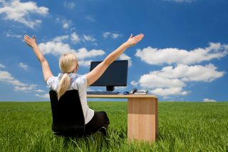 Female teacher sitting at a desk using a computer in a green field raising her arms into a bright blue sky with fluffy white clouds. 