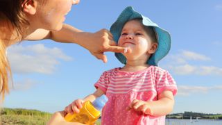 a toddler stands on the beach wearing a blue hat and pink and white striped shirt. An adult woman on the left side of the frame helps to apply sunscreen to the child's face