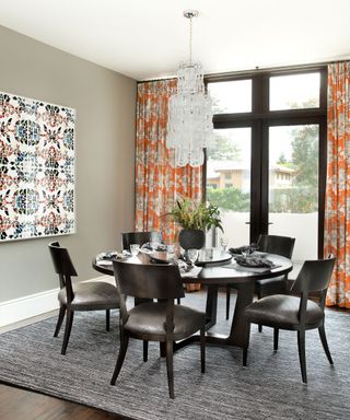 Gray painted dining room with black wooden dining chair, matching dining chairs with leather seats, gray rug, bright orange curtains, large colorful artwork on wall