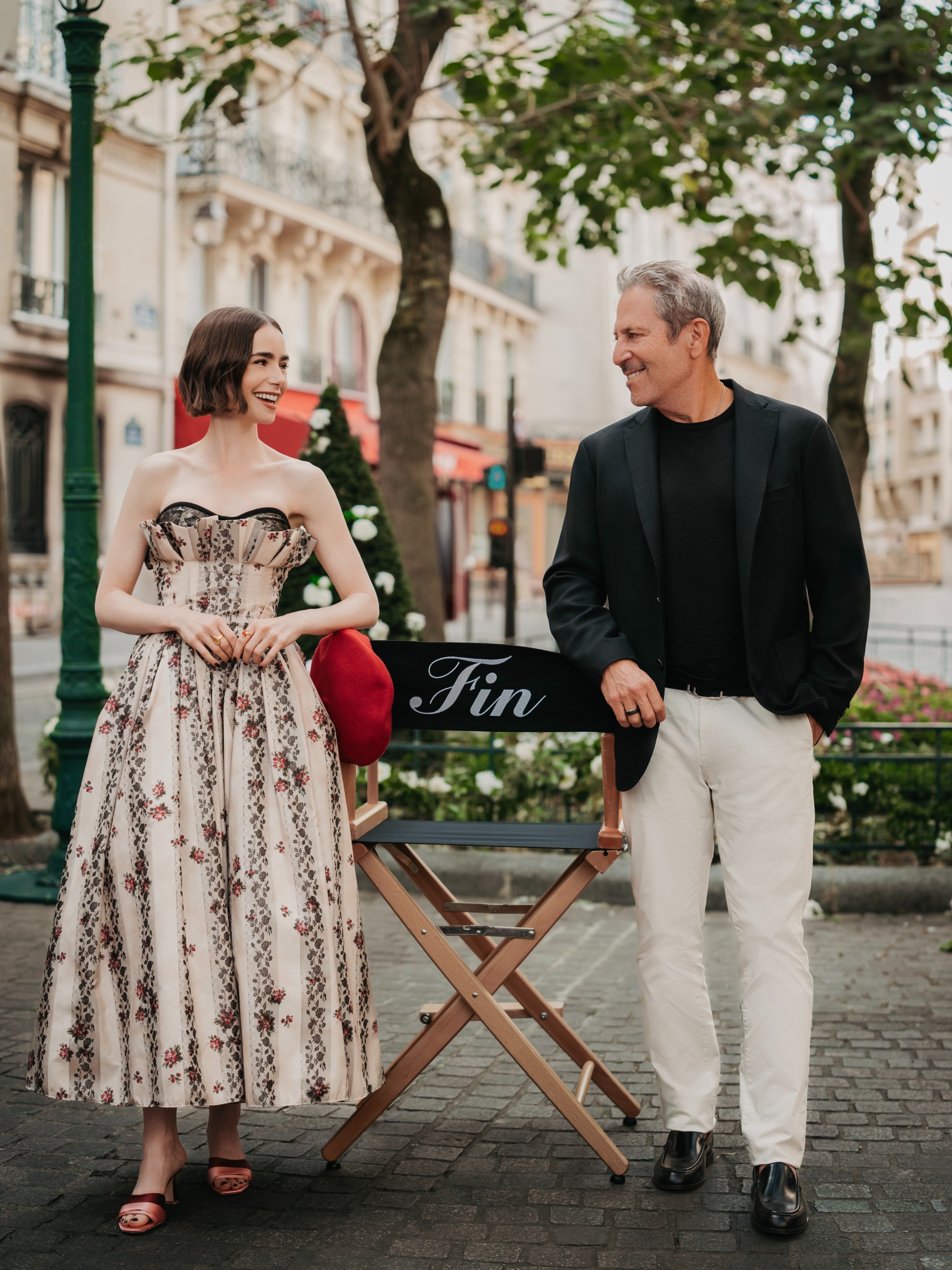 lily collins on set of the last day of emily in paris wearing a floral long dress with her hair in a bop smiling and looking at showrunner darren star