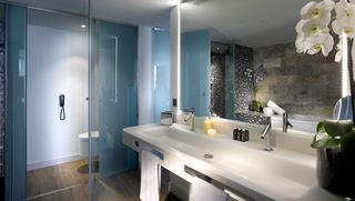 Bathroom image, white sink and units, wooden floor, large mirror above the sink, tinted blue glass wall, white toilet, white flowered plant in a black pot, candles, stone wall reflected in the mirror, ceiling spotlights