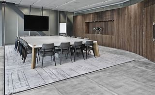 Interior meeting space at the G-Star RAW headquarters, Amsterdam, white top, wooden leg large meeting room table with grey chairs , wooden panel wall to the right, grey panel far wall with a tv screen mounted, white board on silver stand, grey marble effect floor, grey blueprint design square floor design beneath the meeting room table, robot dog in a display box built into the wooden wall