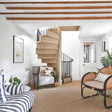 White living room with beige carpets, a stripey sofa on the left, a wicker accent chair on the right, and a spiral staircase at the back of the room