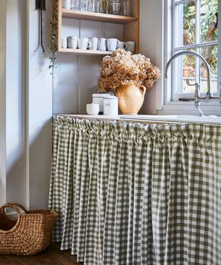 A kitchen sink with a green gingham curtain instead of a cabinet door beneath it