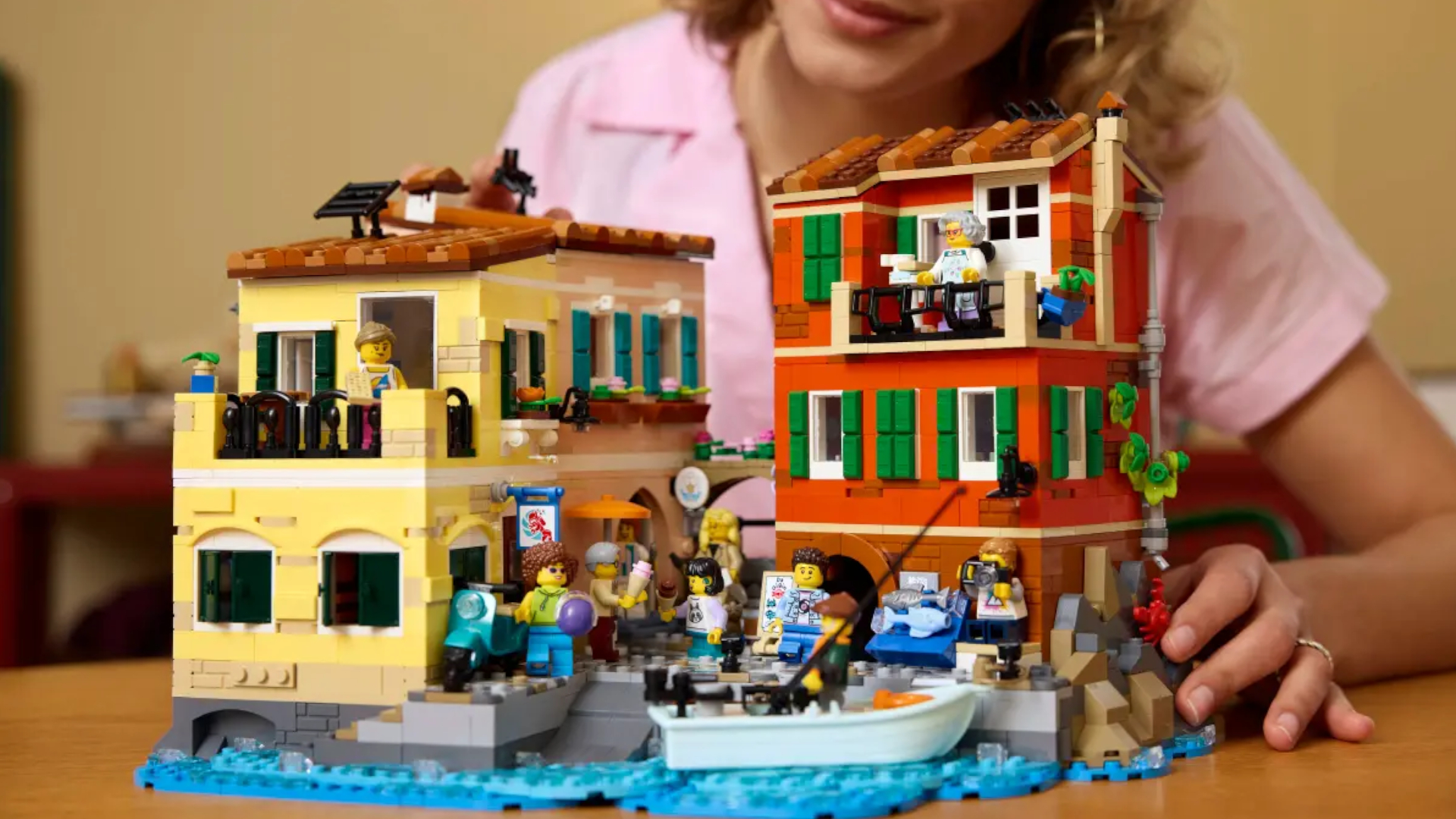 A woman smiles while holding the Lego Italian Riveria model, which sits on a wooden table