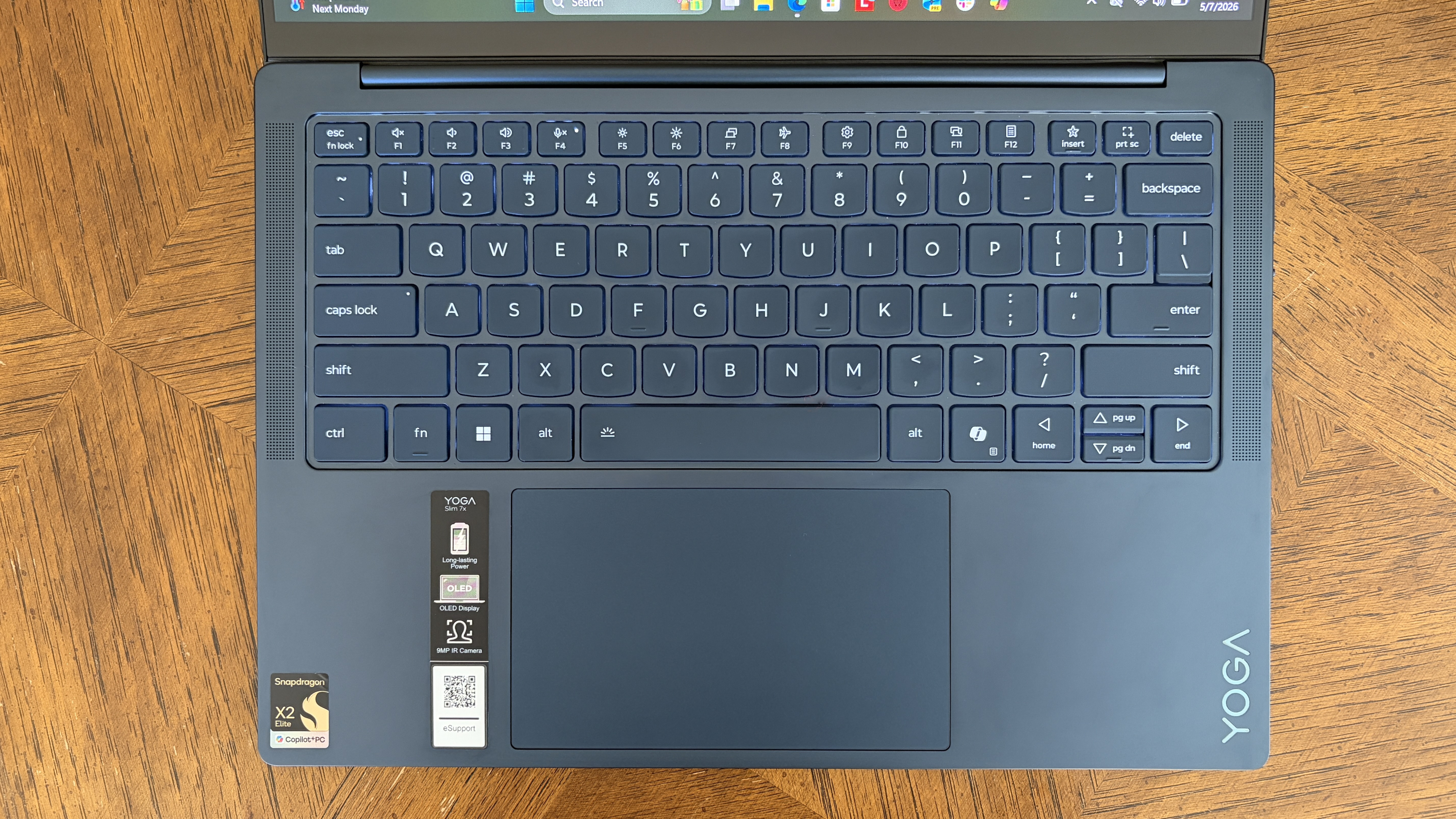A top view of a dark gray Lenovo Yoga laptop keyboard on a wooden table. The keyboard is backlit with white letters, and the trackpad is centered below.