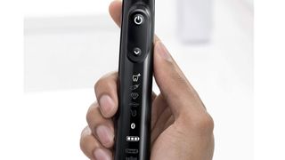 Image shows a hand holding a black Oral B Genius X toothbrush, with a closeup of the backlit indicators on the toothbrush's body.