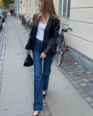 Scandinavian fashion creative Clara Dyrhauge walking on a sidewalk wearing a black leather blazer jacket, white basic scoop neck top, mini bag with a ring handle, straight-leg jeans, and slingback heels.