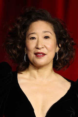 Image of Sandra Oh with a curly brunette bob, wearing a low V-neck black dress and a dark red lipstick