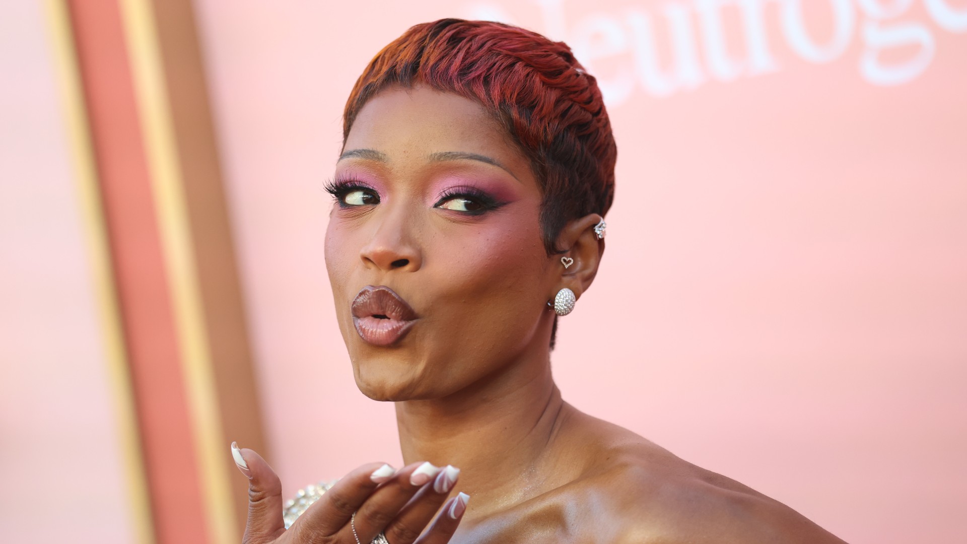 a black woman with short red hair wears pink eyeshadow and poses in front of a peach background
