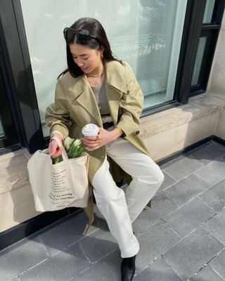 stylish woman sits outside a cafe with a tote bag full of flowers, a coffee in hand, wearing a trench coat, gray t-shirt, white jeans, and black boots