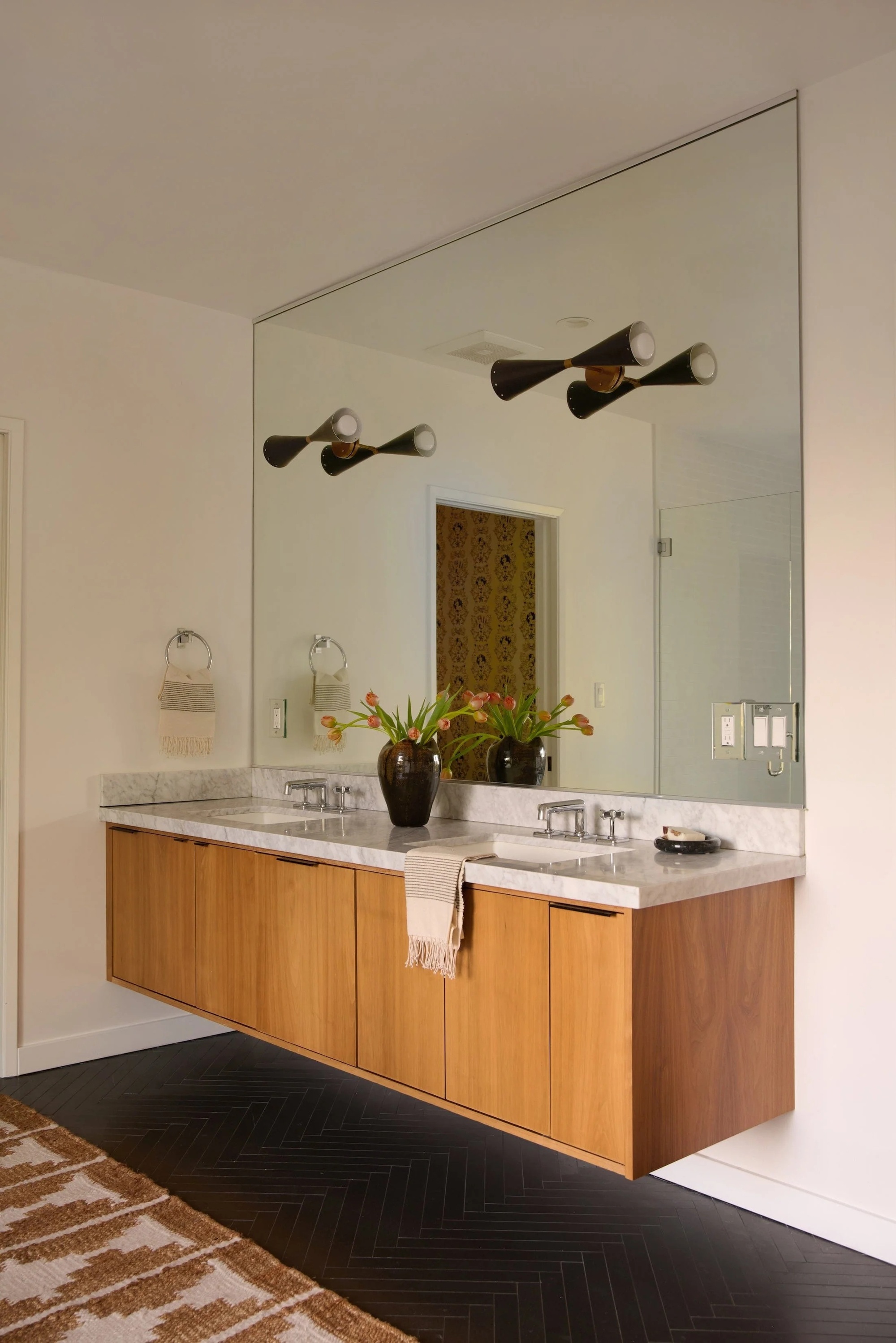 bathroom with large mirror, two wall sconces, floating timber vanity, black herringbone floor tiles and bathroom rug