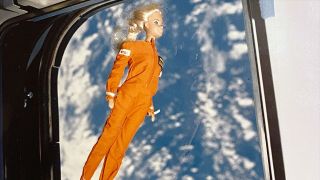 a barbie floating inside a spacecraft in an orange spacesuit. earth is visible out a window behind barbie