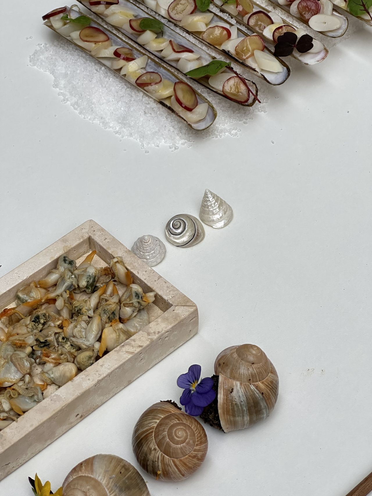 A series of couscous food sculptures, snails, shells, and tomatoes is beautifully positioned on a white cloth table along with silver and wooden cutlery.