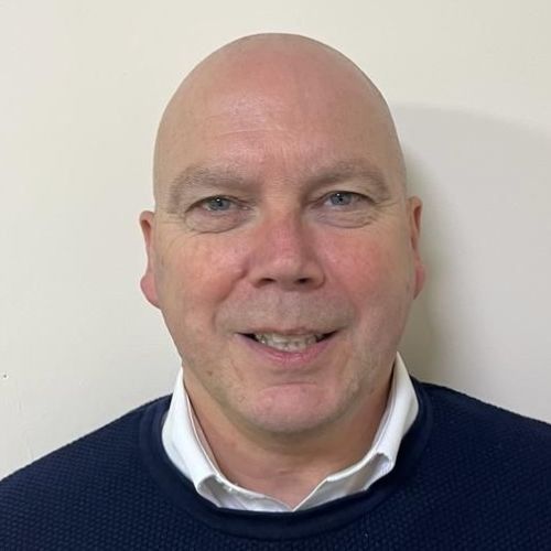 Headshot of Derek Bruce wearing blue sweater and white shirt against white wall background.