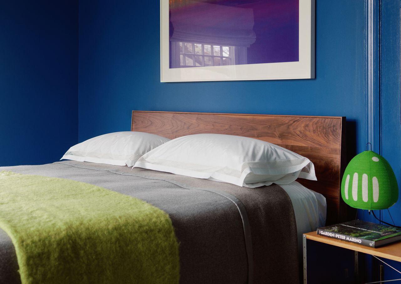 A bedroom painted in a blue Benjamin Moore paint with a purple framed poster on the wall, a green throw on the bed and a neon green table lamp on the bedside table