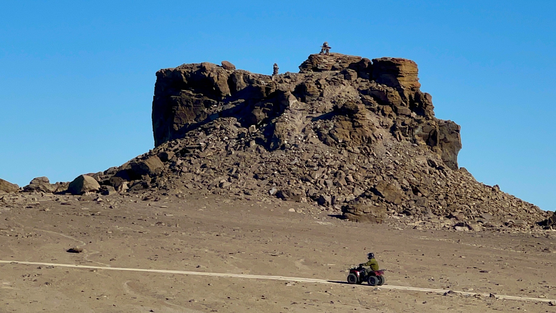 A person on an ATV rides in front of a large, rectangular rock formation with clear blue skies overhead.