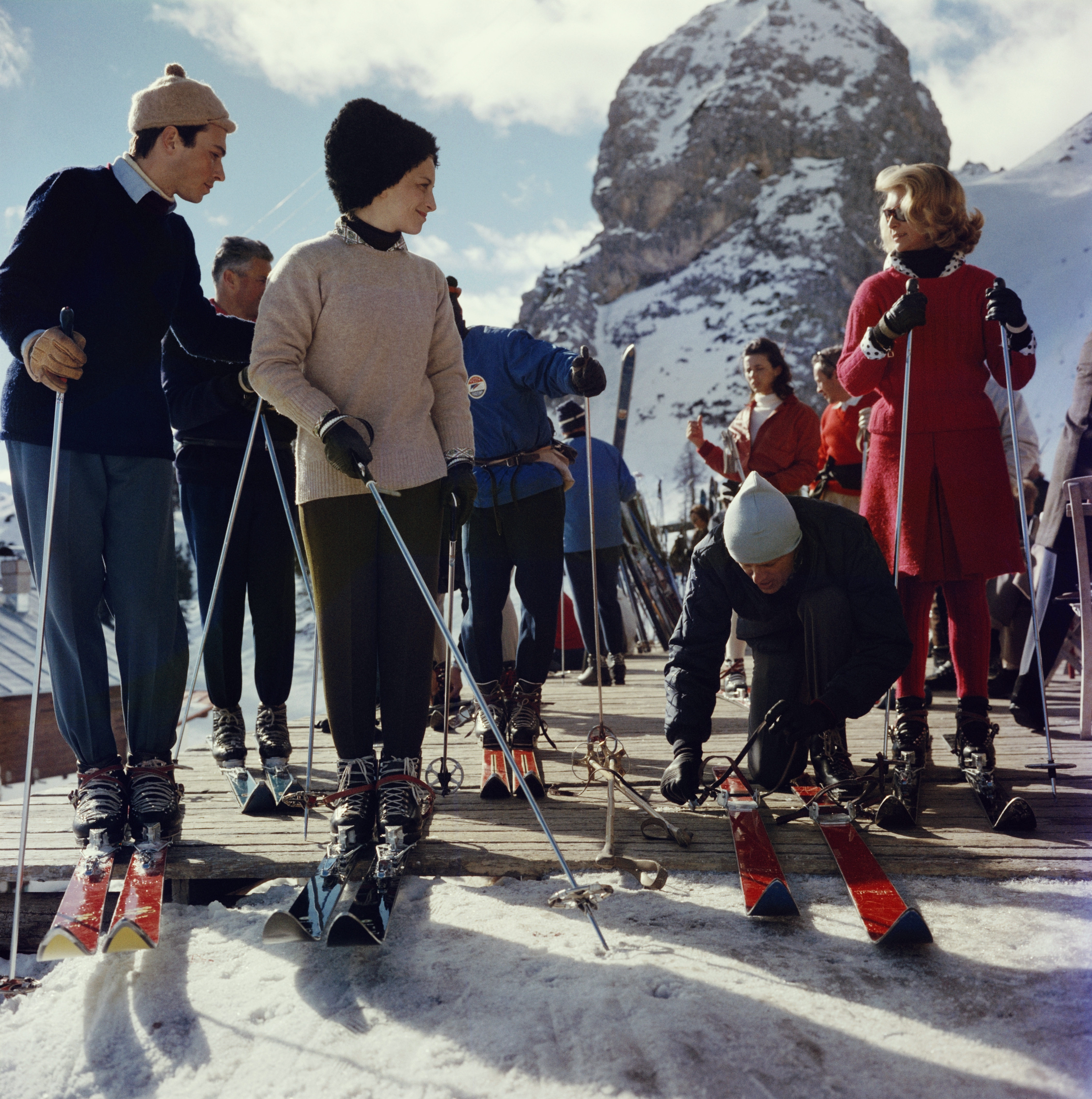 A group of skiers standing on a wooden platform, adjusting bindings and holding ski poles, dressed in knitwear and winter hats with snowy peaks rising behind them.