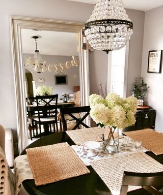A small farmhouse dining room with a black circular table with jute placemats and white flowers, a beaded chandelier above it, and a tall distressed white mirror behind it reflecting the scene