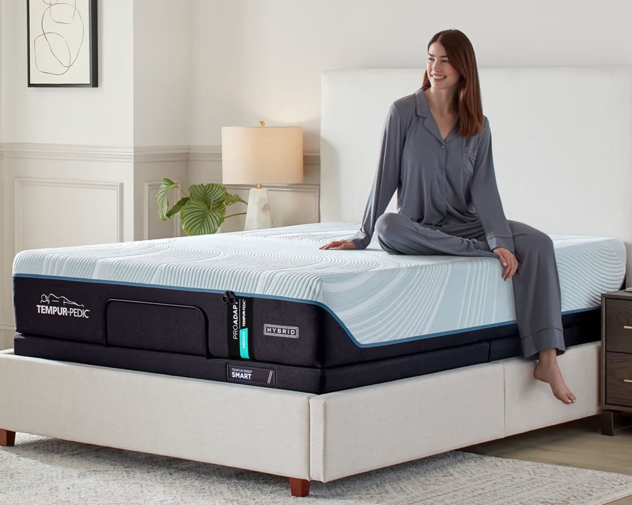 A woman with brown hair in grey pyjamas sitting on the Tempur-Pedic Pro-Adapt mattress in a white, minimalist bedroom with panelled walls and a large pale grey rug on the wooden flooring.