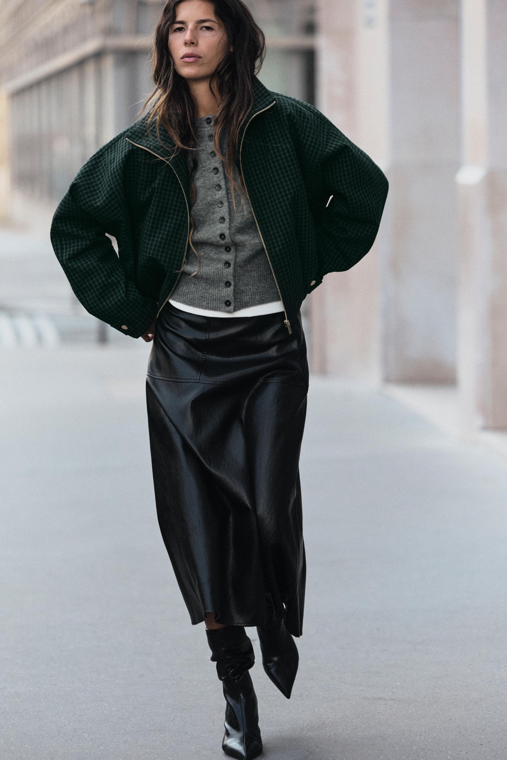 Model wears green plaid bomber, gray cardigan, white t-shirt, black leather skirt, and black slouchy boots.