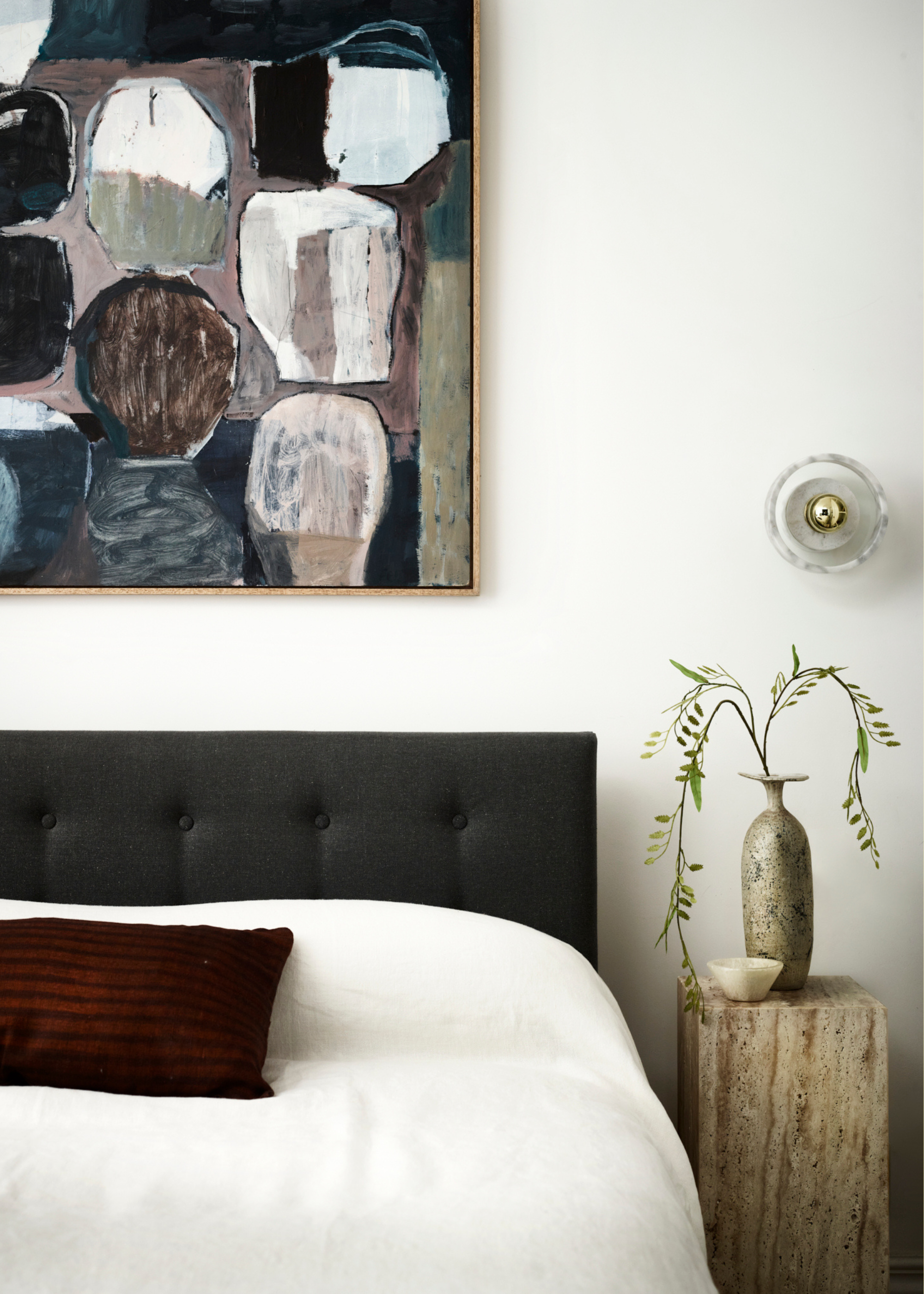 A white bedroom with a framed painting above a black bed with white bedding and a red striped accent pillow beside a stone side table with a vase and a candle