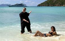 Kim DotCom posing on beach with woman laying in water in a bikini