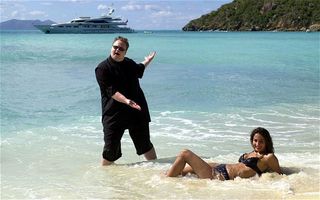 Kim DotCom posing on beach with woman laying in water in a bikini