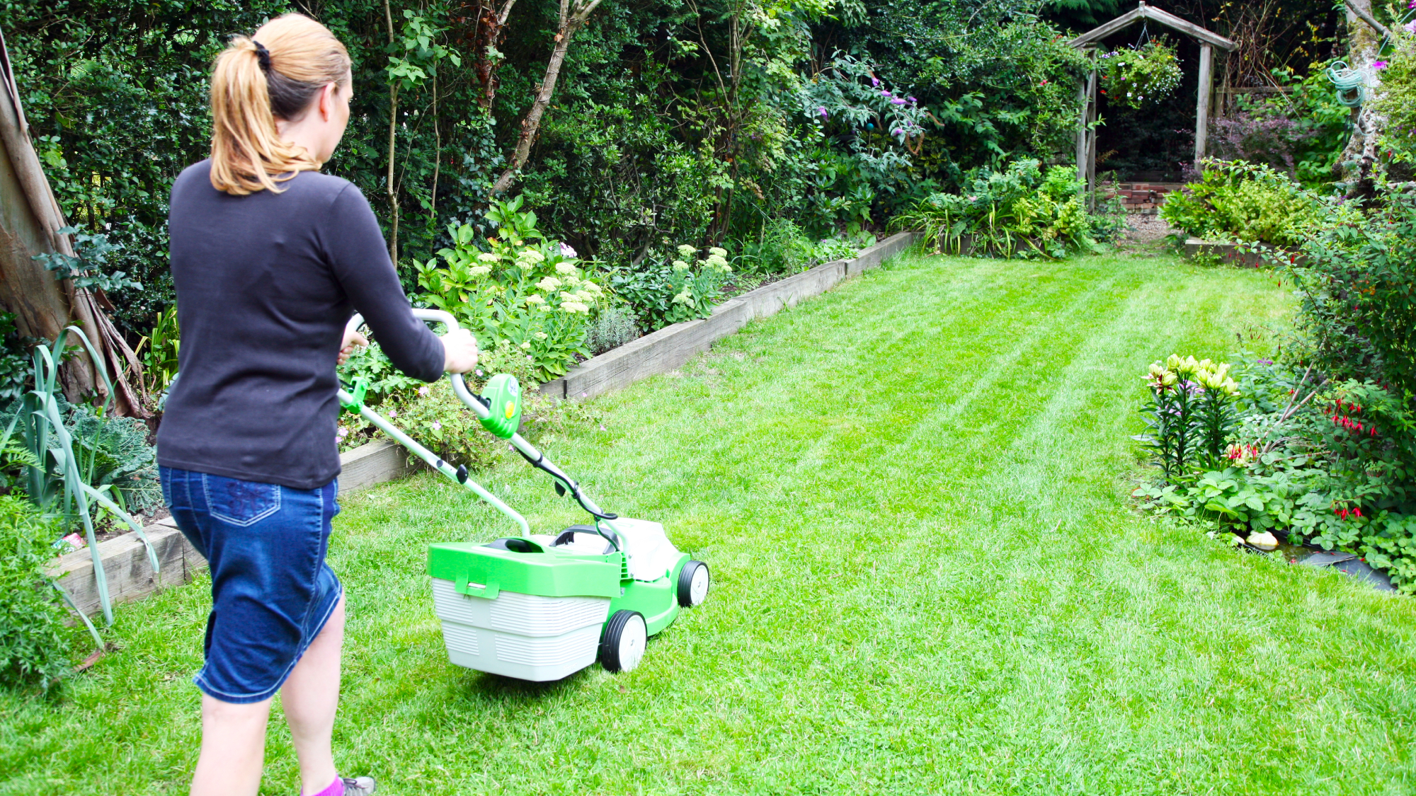 young woman mowing a healthy looking lawn in summer using a lawnmower as part of a good lawn care routine