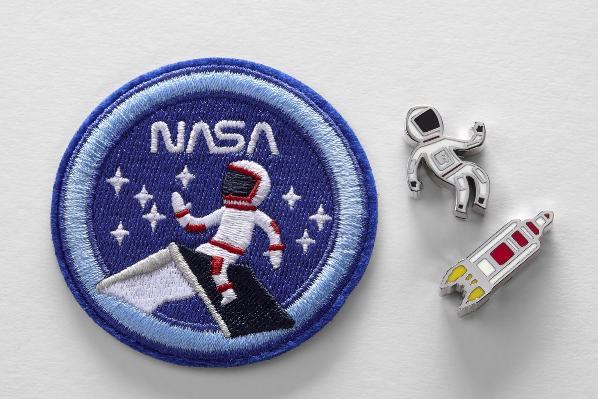 Embroidered patch and 2 pins from the NASA-inspired Limited Edition Notebooks from Moleskine. The patch is a round dark blue patch with a light blue border. The word "NASA" is at the top in white. A person in a white astronaut suit with red trim is sitting on the roof of a house. They are surrounded by white stars. The pins are a small person in a white astronaut suit with red trim and a small grey, white, and red simplistic rocket blasting off.