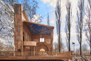 Babyn Yar Synagogue is set in a park, surrounded by tall trees with naked branches. The Synagogue sits on a wooden platform and is made entirely out of wood with a steel structure. The interior is painted with Jewish religious symbols.