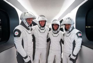 Four astronauts in white and black spacesuits stand inside a white-walled corridor.