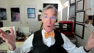 Bill Nye the Science Guy, wearing a yellow bow tie, white shirt and vest, gestures while sitting in his office.