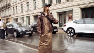 A guest wearing a brown oversized leather jacket, light brown maxi dress, black bag, black cap, and sunglasses outside A. Roege Hove during the Copenhagen Fashion Week Spring/Summer 2024 on August 7, 2023 in Copenhagen, Denmark.
