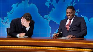 Colin Jost looks into his hands as Michael Che one-ups him during 2026 Weekend Update segment. Both men have on suits and are at the desk. 