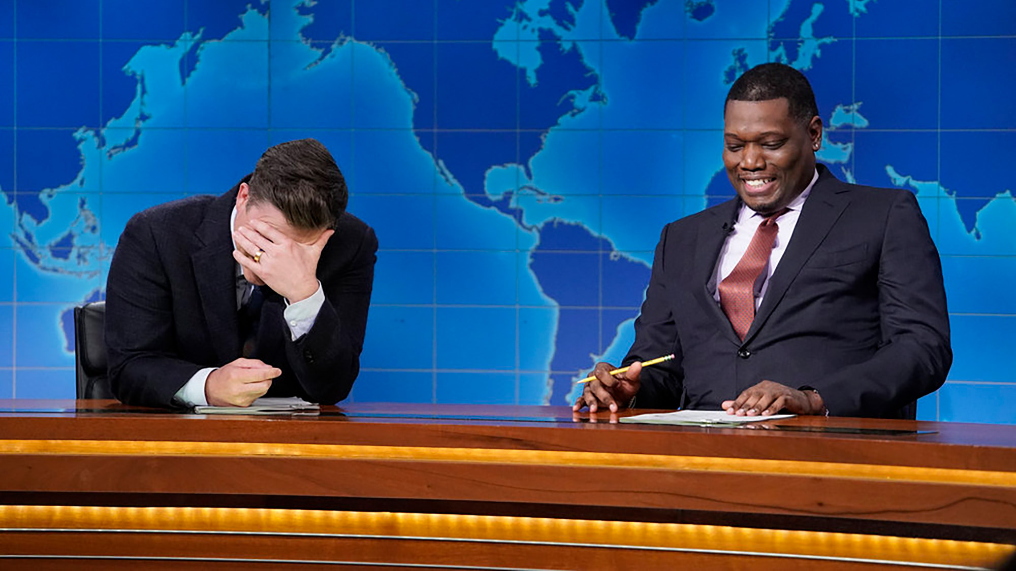 Colin Jost looks into his hands as Michael Che one-ups him during 2026 Weekend Update segment. Both men have on suits and are at the desk. 