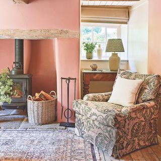 armchair next to inglenook fireplace with woodburner in small Cotswold cottage, with pink painted walls and rug on bare floorboards