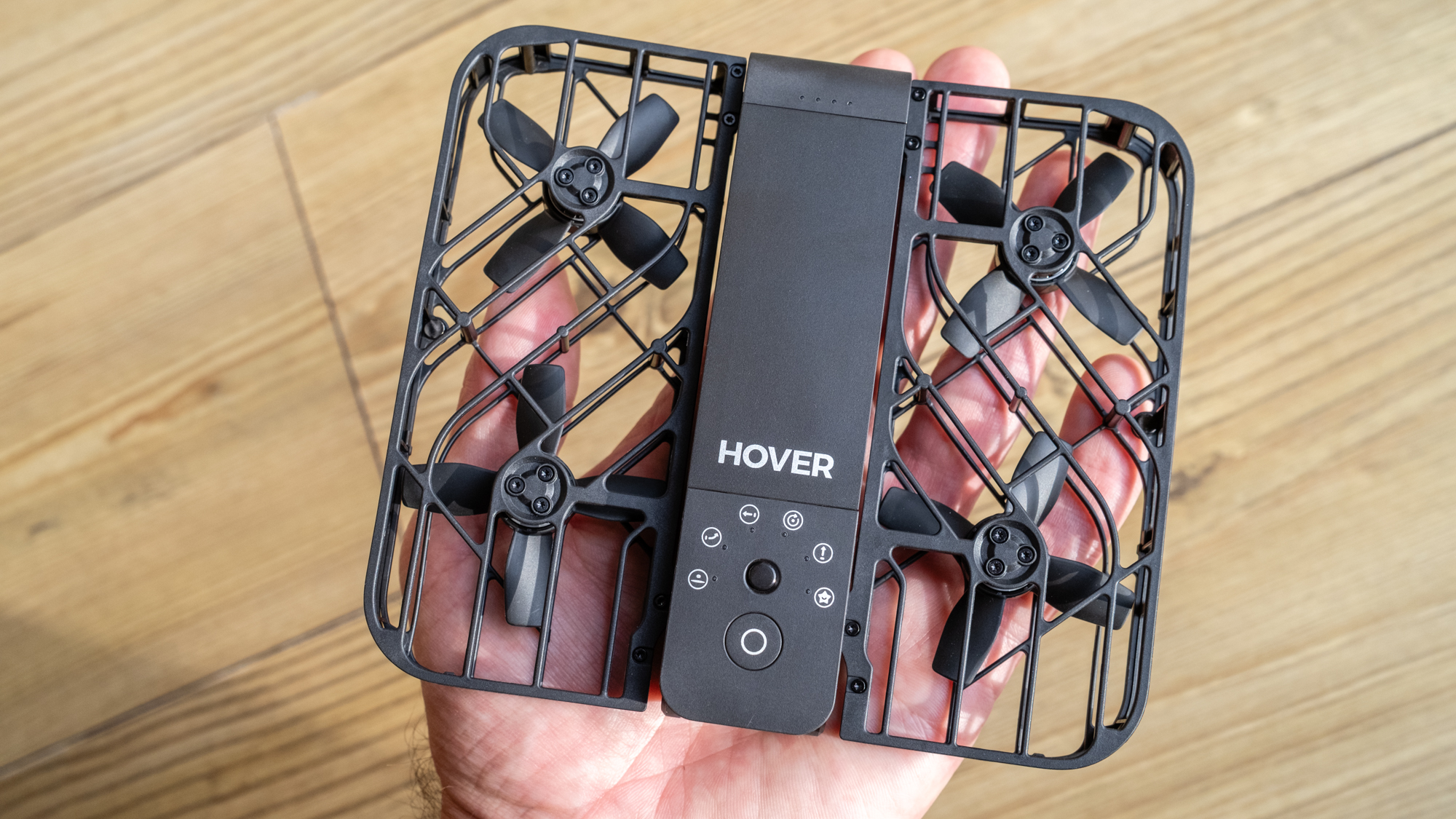 a square drone sits in someone's hand. the quad copter blades are protected on top and bottom by a black grid.