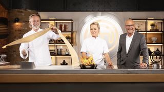 Marcus Wareing, Anna Haugh in chefs' whites and Gregg Wallace in a dark suit and white shirt stand at a kitchen counter as Marcus cuts pasta and Anna uses a frying pan in MasterChef: The Professionals.