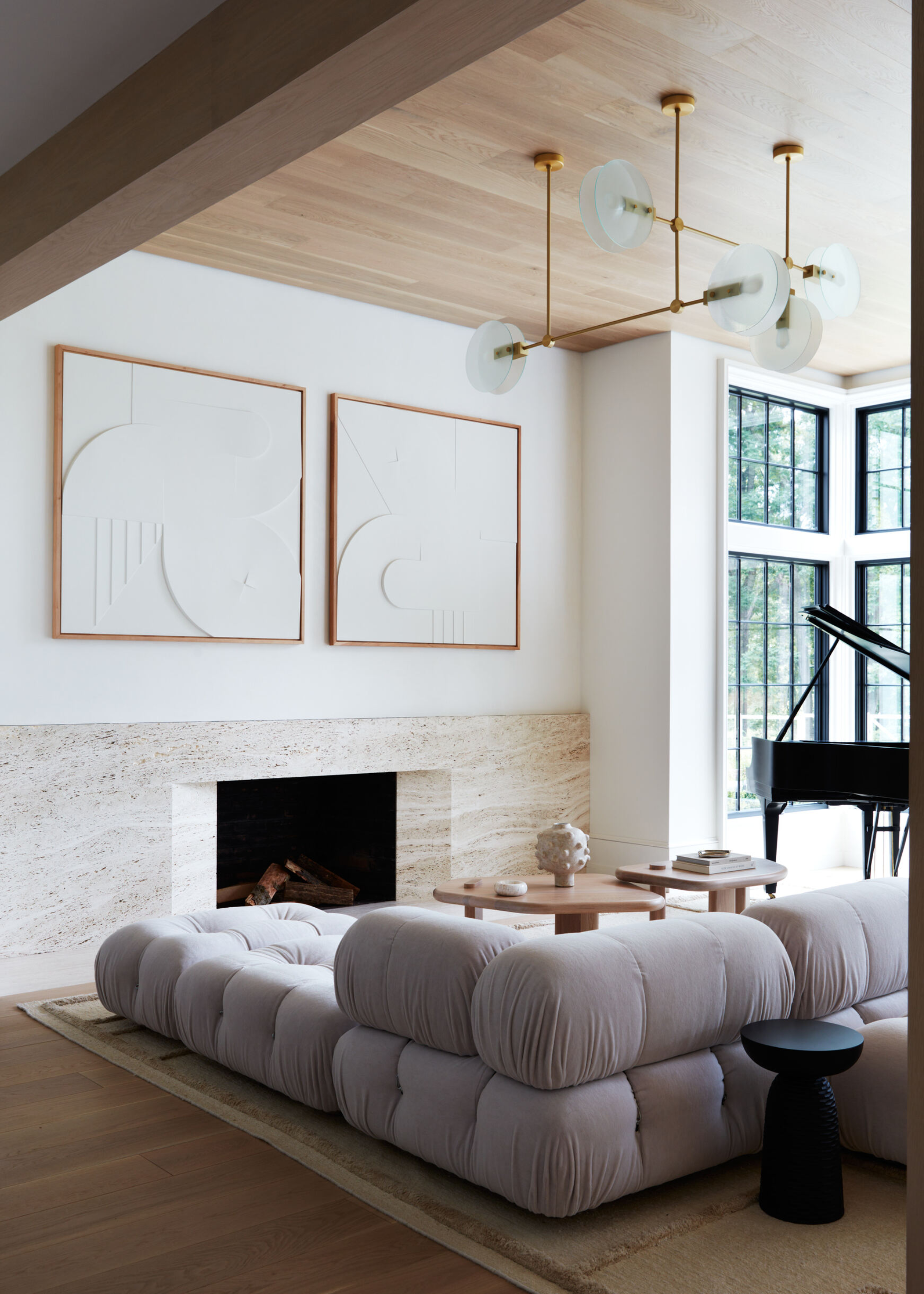 beige living room with travertine slab fireplace, two sculptural white artworks hung above, a timber clad ceiling, pendant light, piano in the corner surrounded by steel-framed windows, and a B&B camaleonda sofa