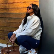 Influencer @jastookes crouches down wearing navy leggings with white New Balance trainers, a white fleece and a brown Hermes bag.