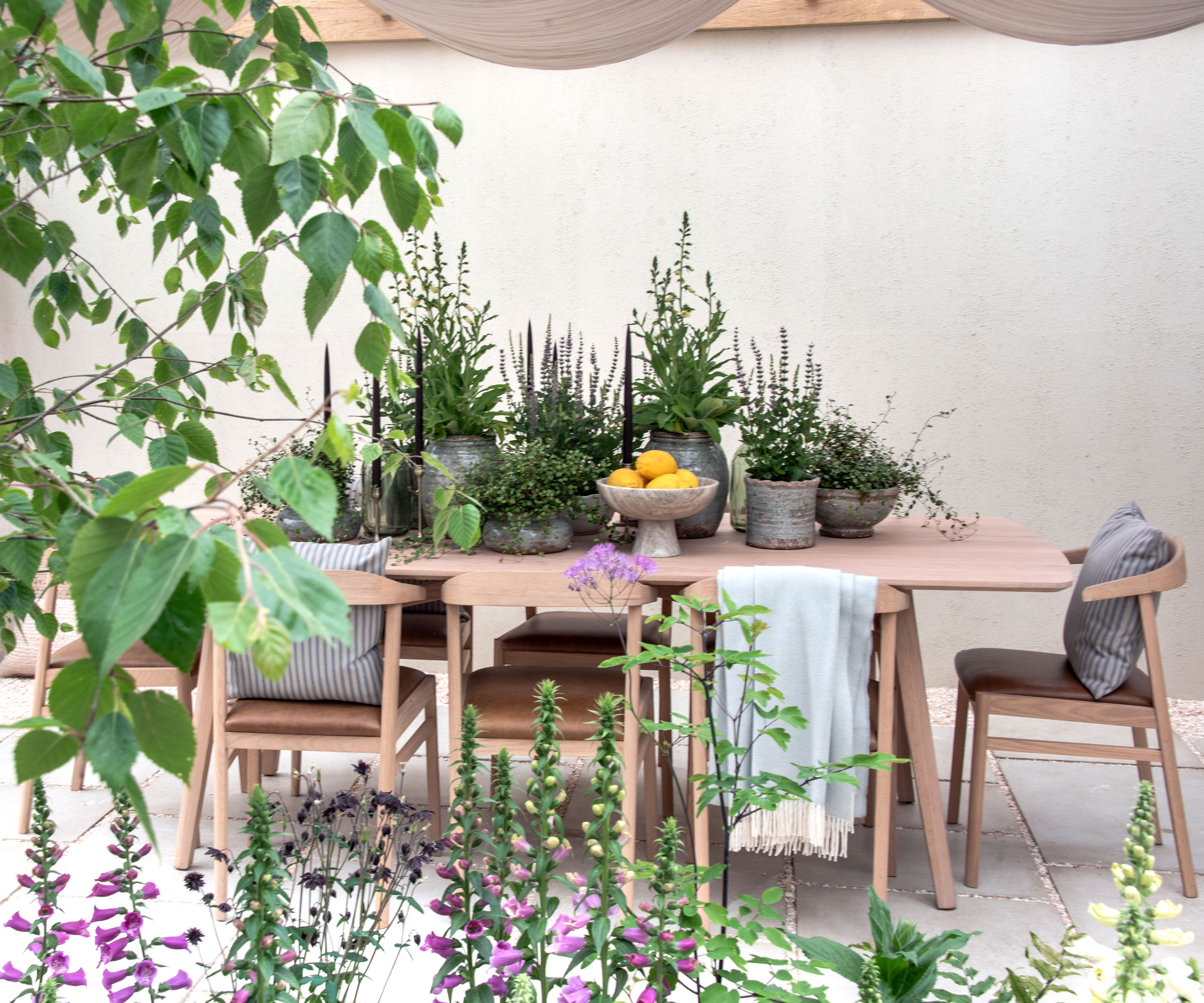 garden terrace with outdoor dining tables and chairs, with accessories such as cushions and throws, pot plants and candlesticks, and foxgloves planted in a flower bed