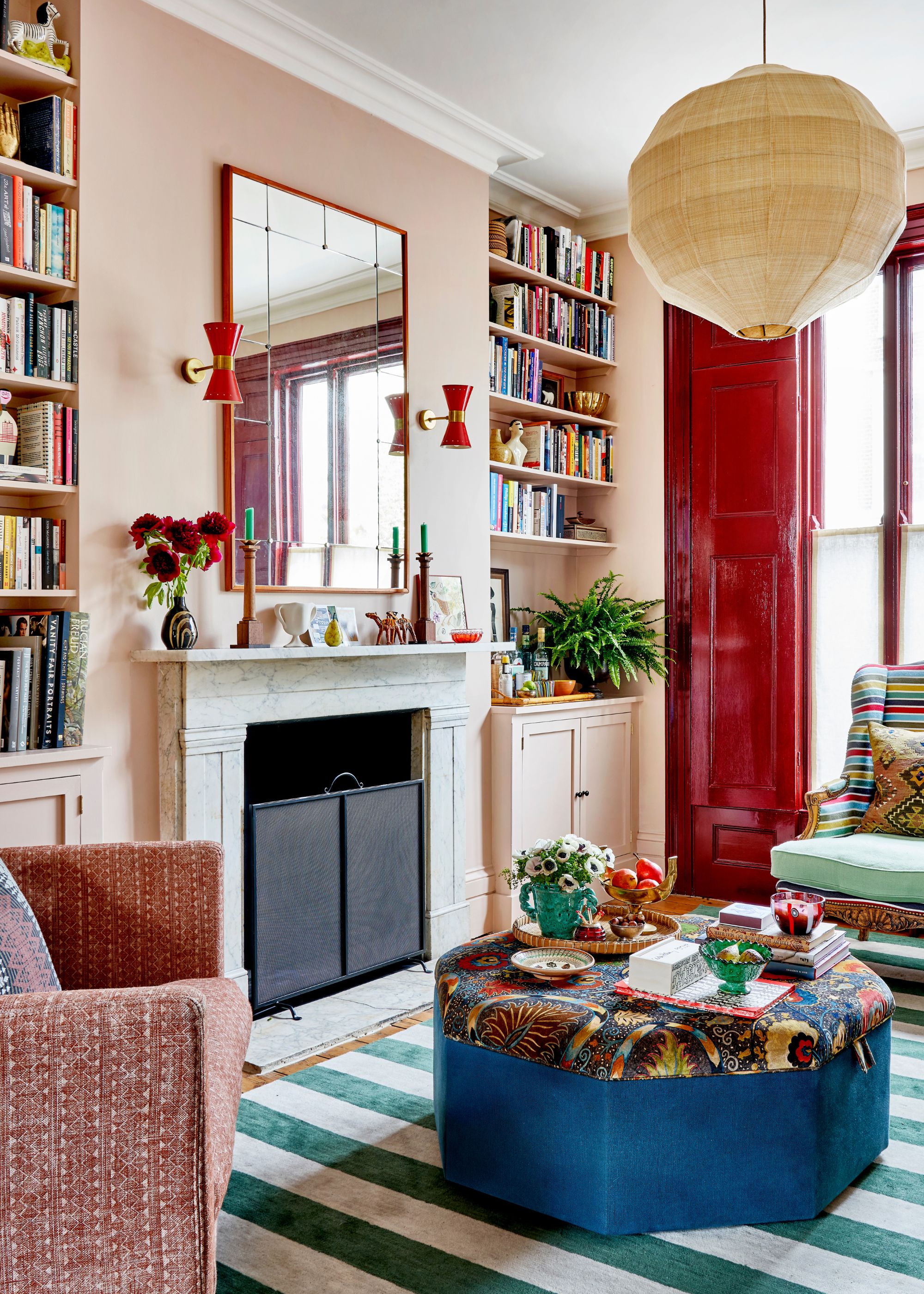 A living room with plaster pink walls, a dark red bay window, a green striped rug, a fireplace with a red mirror and two red wall lights, and built-in bookshelves.