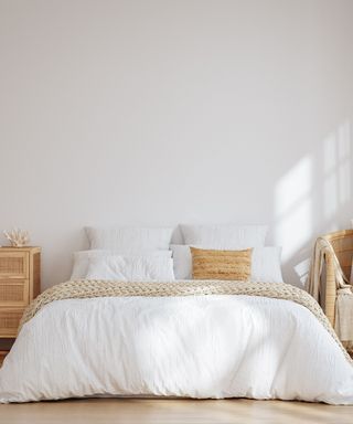 A bedroom with white walls, a bed with white sheets and beige throw pillows and bed runners, a wooden nightstand and chair, and a wooden floor