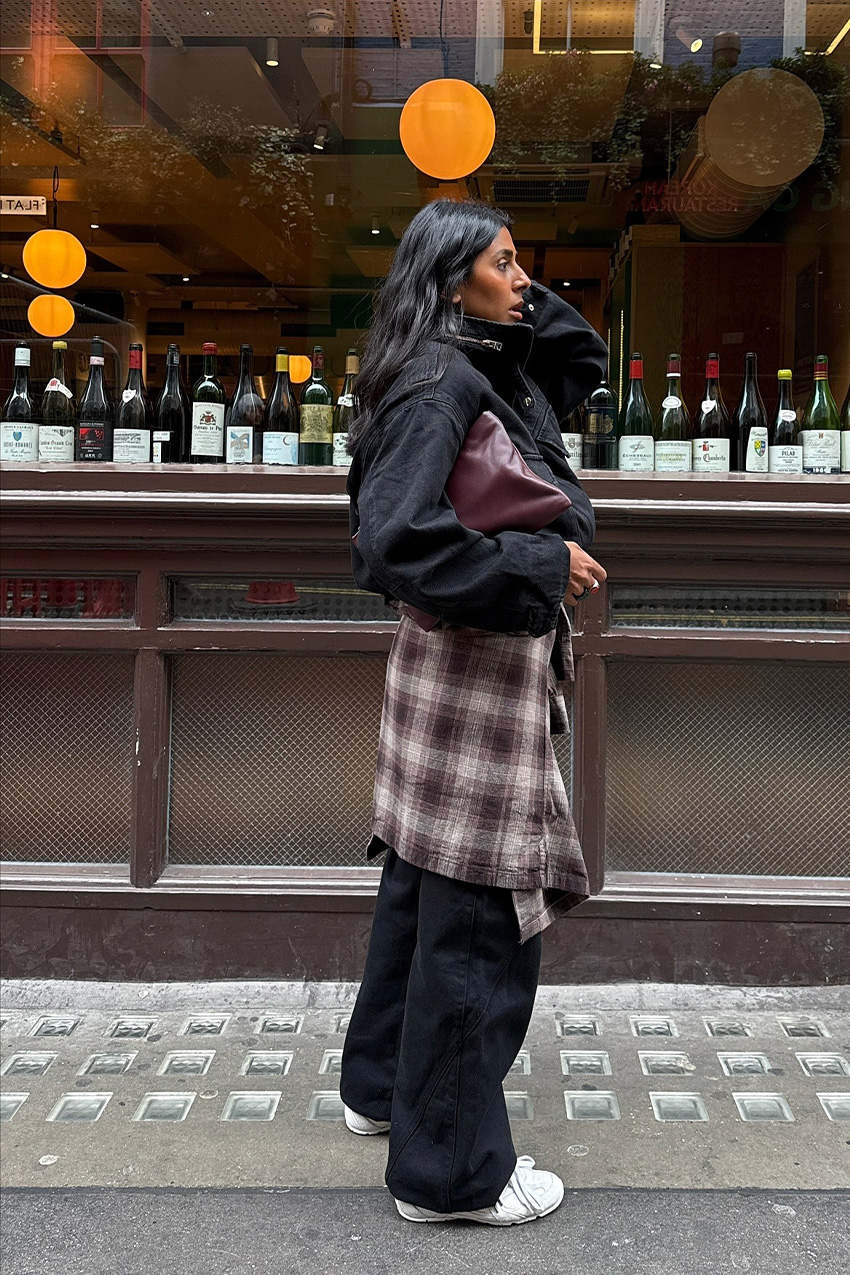 A woman standing on the sidewalk in front of a restaurant in London wearing a black funnel neck bomber jacket, black baggy wide leg jeans, a brown plaid flannel tied around the waist, a burgundy clutch bag, and white retro sneakers.
