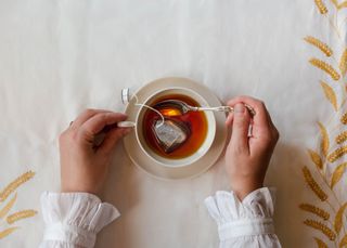 A person holding a tea cup full of tea and a tea bag, while stirring it.